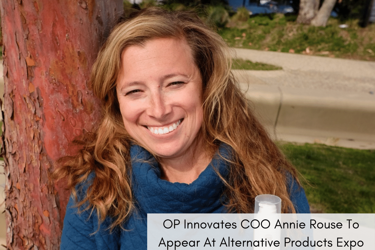 Annie Rouse, a white woman with brown hair in a blue sweater, smiles with arms crossed. She's holding an Overcome Every Day CBD product in one hand. An added headline reads, "OP Innovates COO Annie Rouse Appears At Alternative Products Expo"